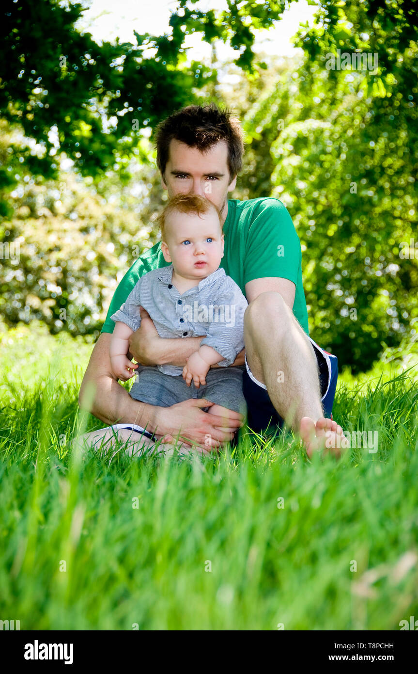 Young dad with baby boy, sitting outdoors in garden on a sunny day. Dad looking at camera. Portrait format Stock Photo