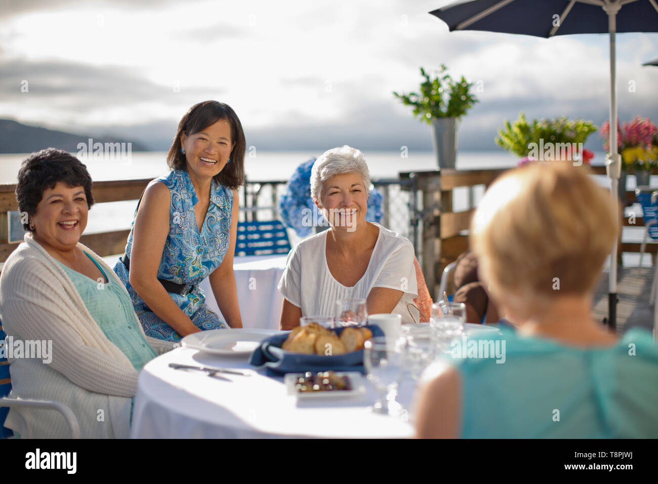 Two mature female friends out to lunch at a seaside restaurant smile and share a laugh with some acquaintances who have come to say hello. Stock Photo