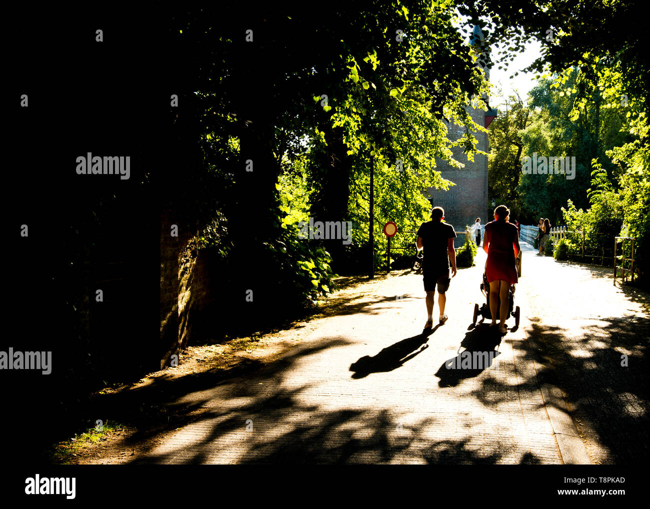 backlit image of young couple in silhouette, walking from dark shaded path, moving forward into sunlight pathway in front of them,pushing a pushchair. Stock Photo