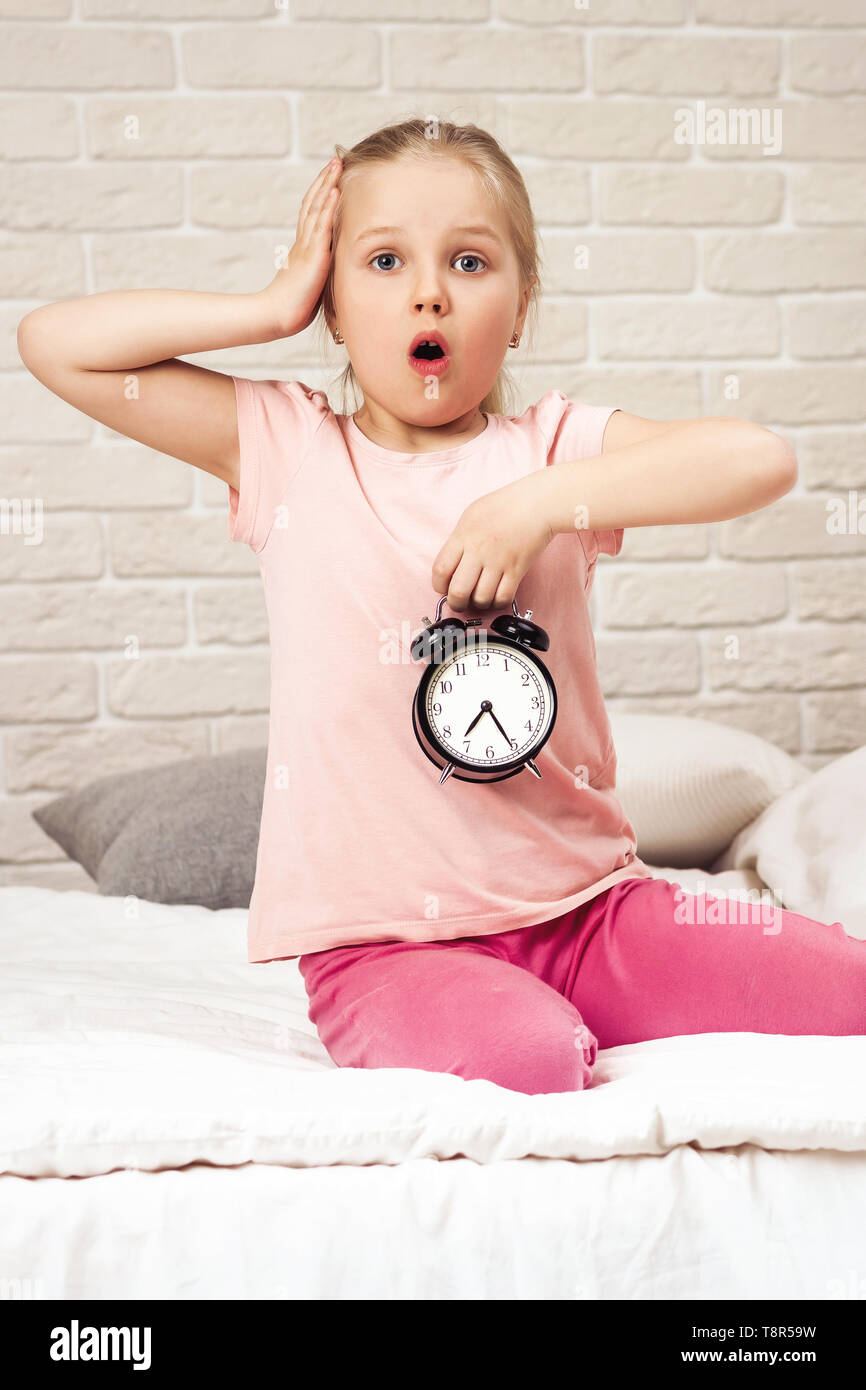 Cute Surprised Little Child Girl In Pyjamas With Clock In Bed