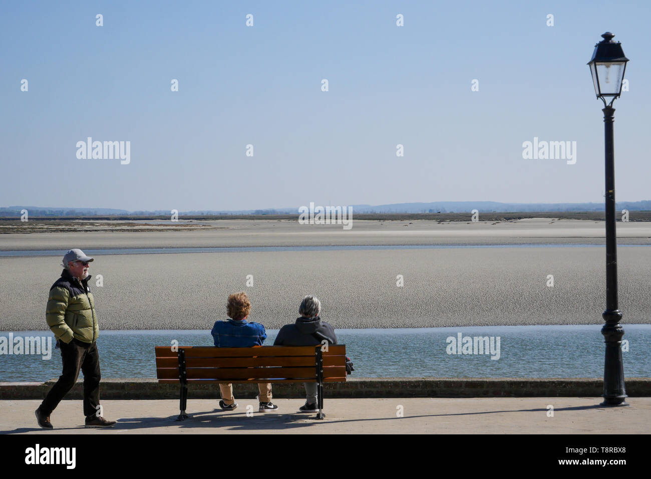 Couple Sitting On A Bench Facing The Seascape At Low Tide Le