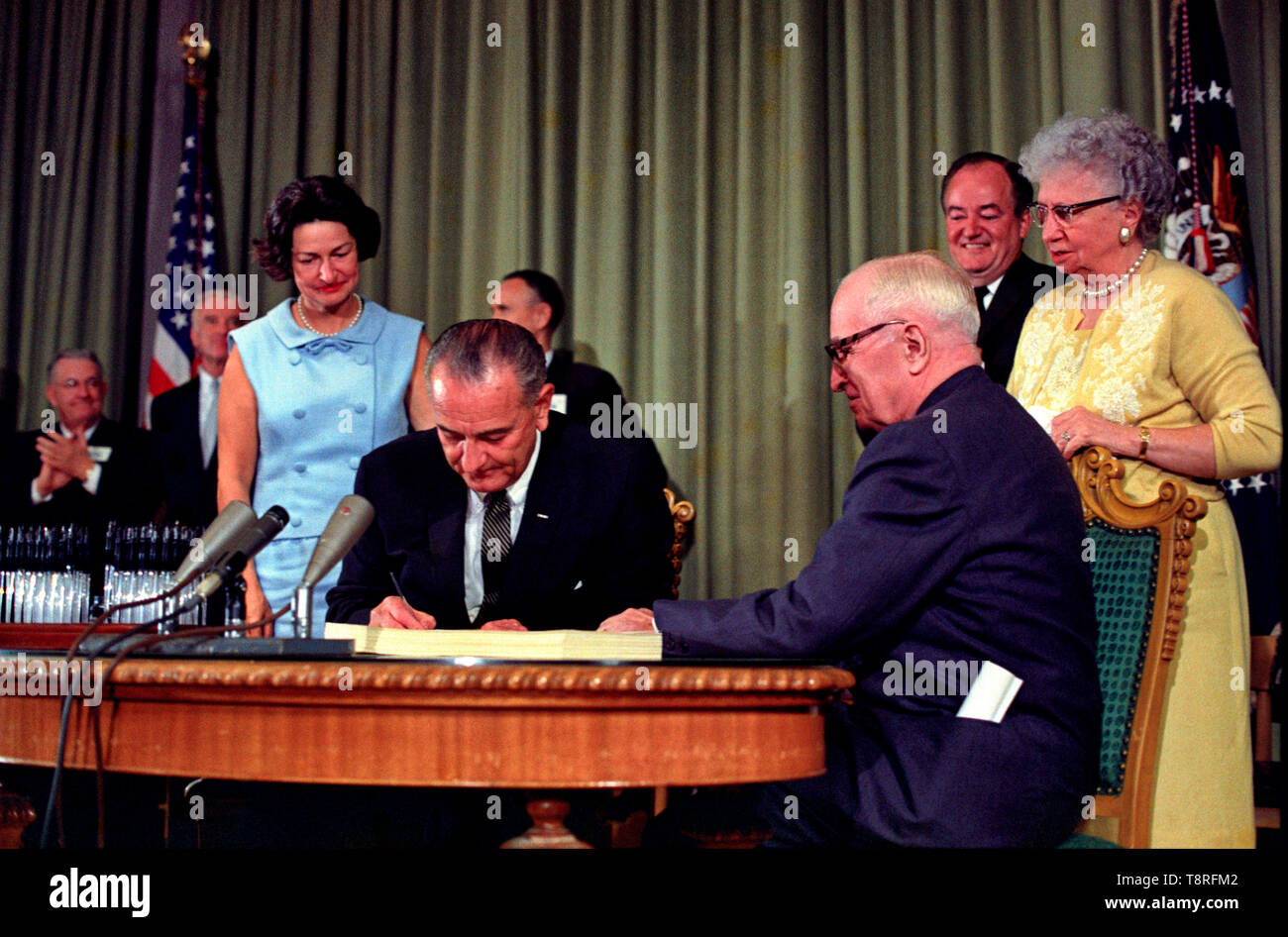 President Lyndon B. Johnson signing the Medicare Bill at the Harry S. Truman Library in Independence, Missouri. Former president Harry S. Truman is seated at the table with President Johnson. The following are in the background (from left to right): Senator Edward V. Long, an unidentified man, Lady Bird Johnson, Senator Mike Mansfield, Vice President Hubert Humphrey, and Bess Truman. July 30,1965 Stock Photo