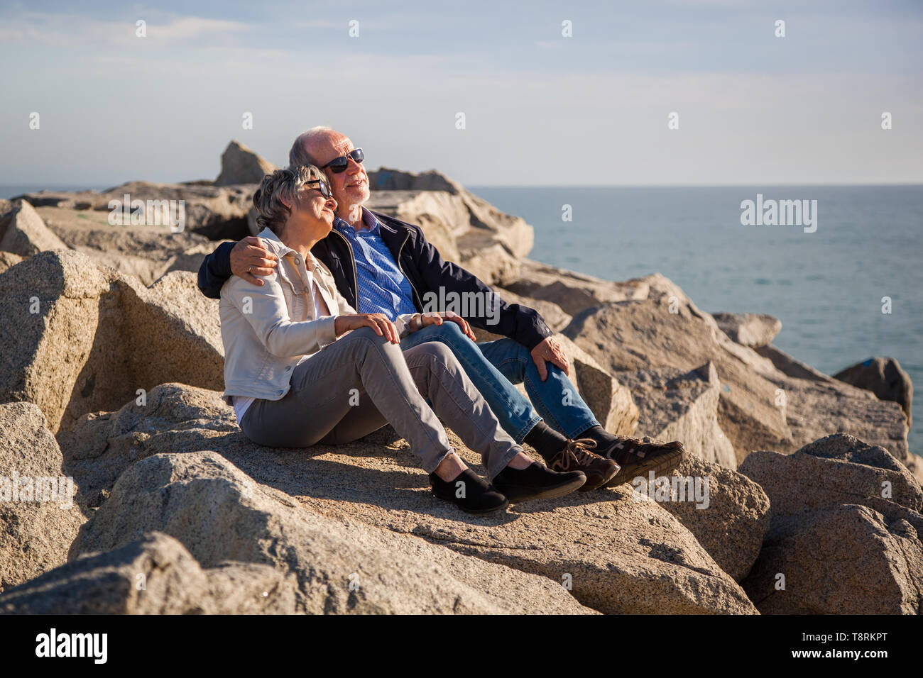 Elderly couple sitting together on sunny rocks by the sea smiling and embracing, concept happy active seniors Stock Photo