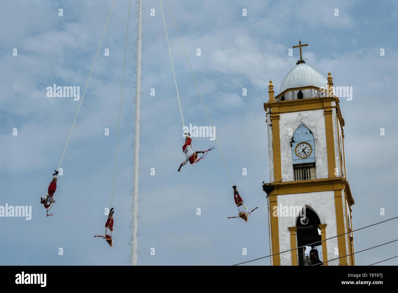 Voladores performs in front of the Church of the Assumption in Papantla, Veracruz, Mexico. The Danza de los Voladores is a indigenous Totonac ceremony involving five participants who climb a thirty-meter pole. Four of these tie ropes around their waists and wind the other end around the top of the pole in order to descend to the ground. The fifth participant stays at the top of the pole, playing a flute and a small drum. The ceremony has been inscribed as a Masterpiece of the Oral and Intangible Heritage of Humanity by UNESCO. Stock Photo