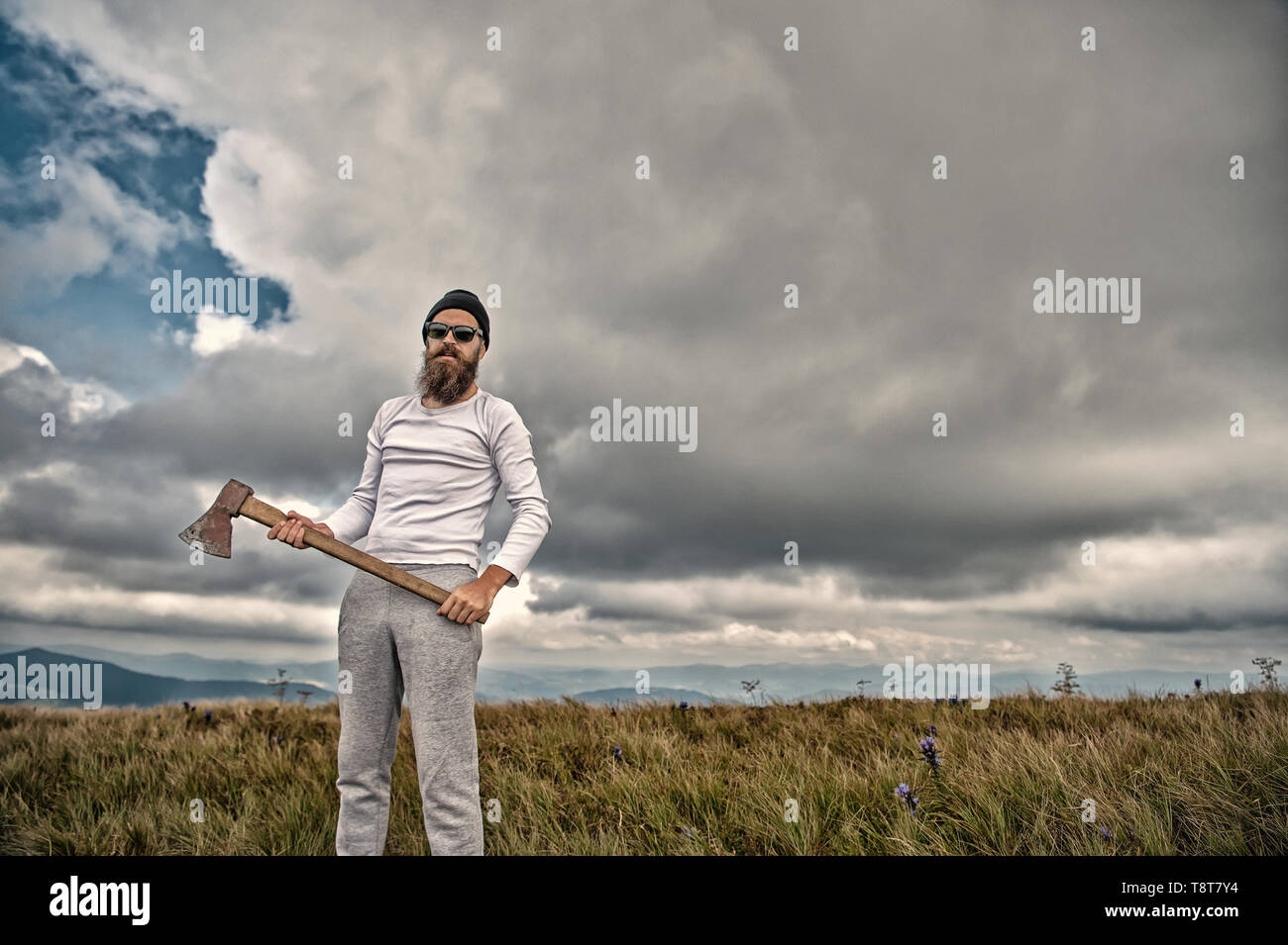 Hipster bearded man in sunglasses with axe stand on mountain landscape on cloudy sky. Logging and chopping concept. Wanderlust, vacation, travelling, vintage filter Stock Photo
