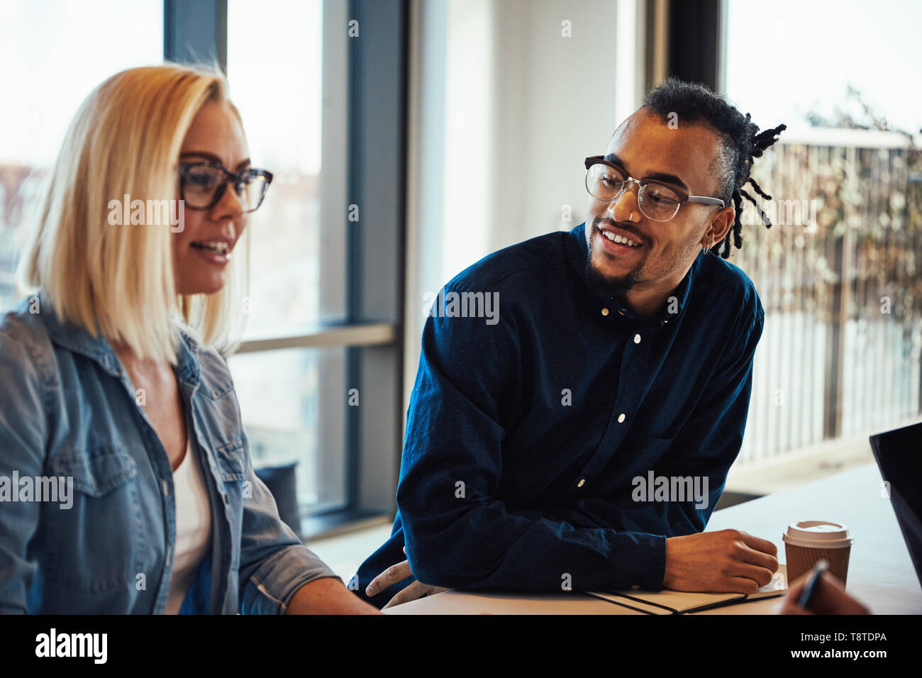 Two diverse young business colleagues smiling while having a meeting around a desk in a modern office Stock Photo
