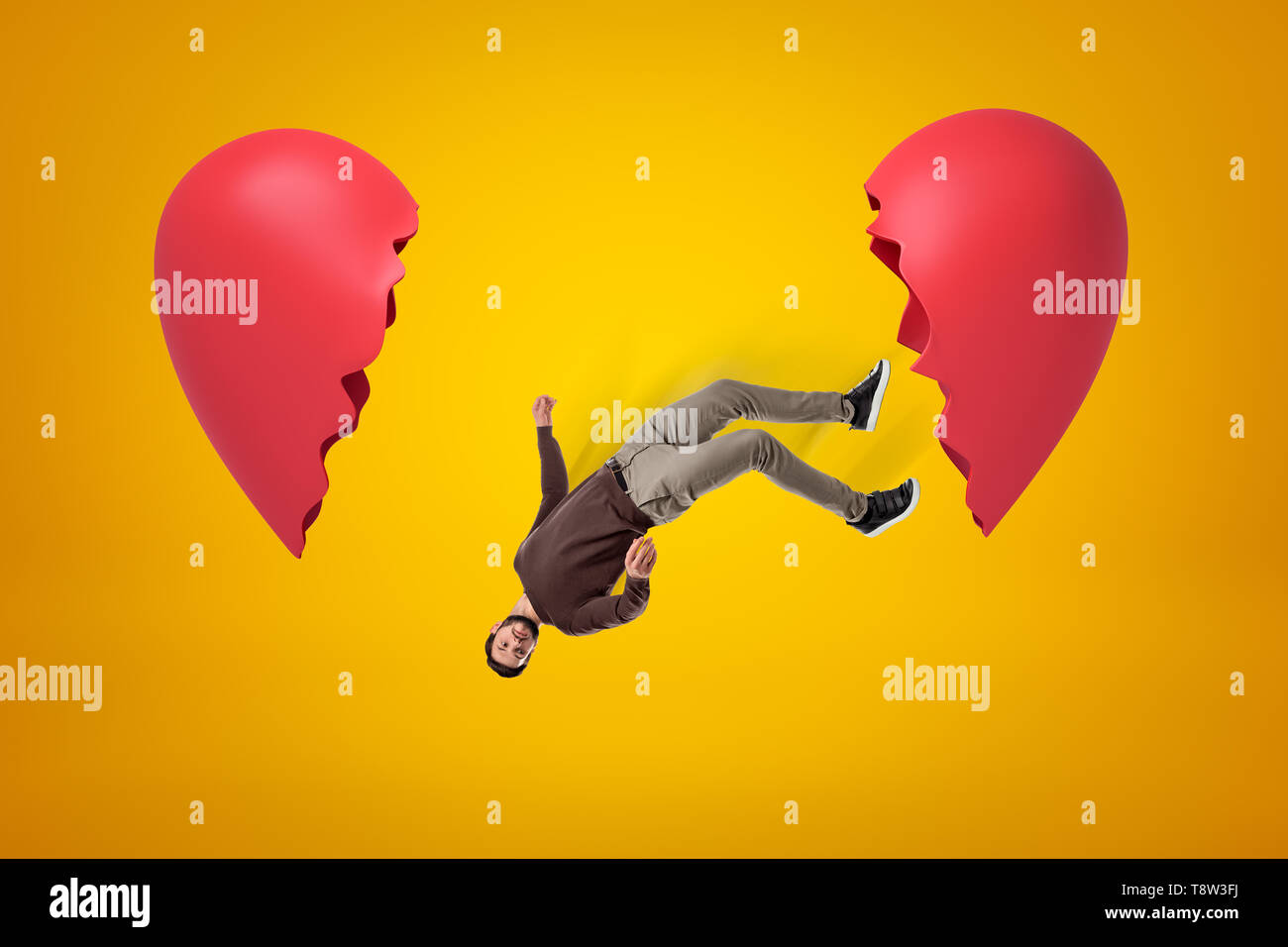 Young man in casual clothes falling between two broken heart pieces on yellow background Stock Photo
