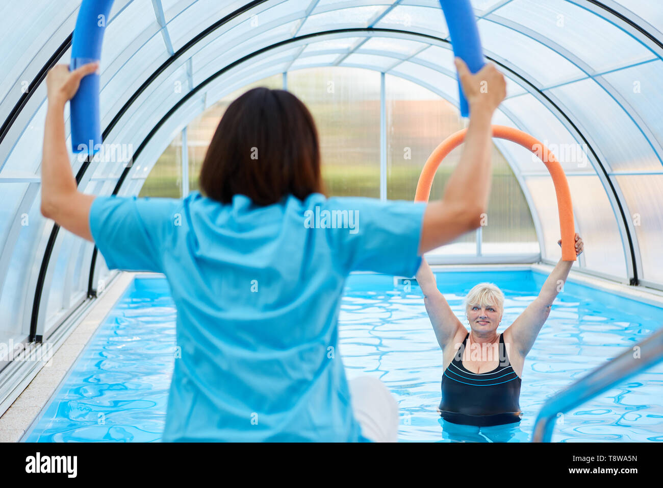 Therapist with swimming noodle shows a senior woman doing aquagym exercise Stock Photo