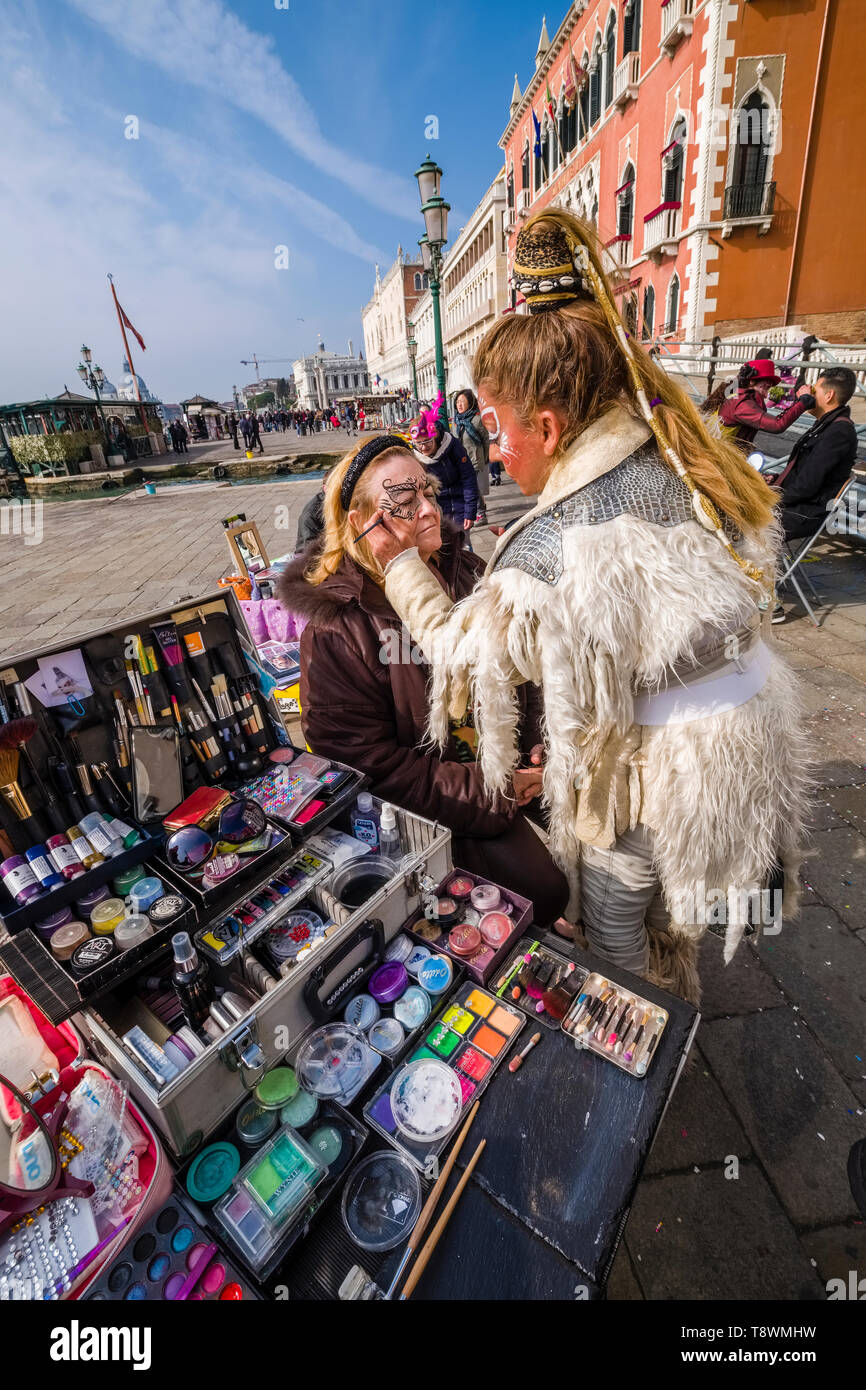 A costumed woman is face painting a child at Grand Canal, Canal Grande, celebrating the Venetian Carnival Stock Photo