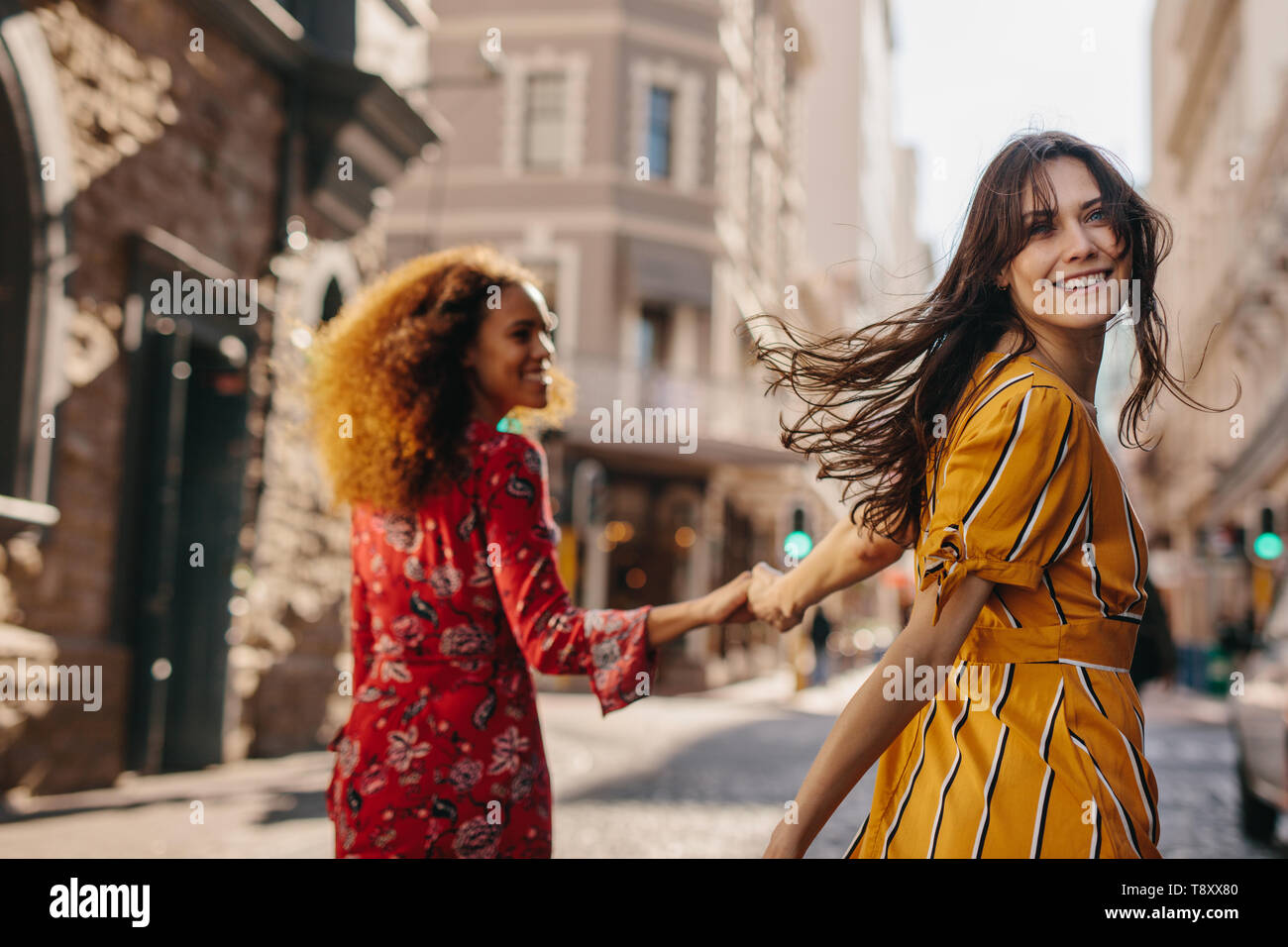 Rear view of two young women walking together on city street holding hands. Woman looking over her shoulder while walking with her friend outdoors. Stock Photo