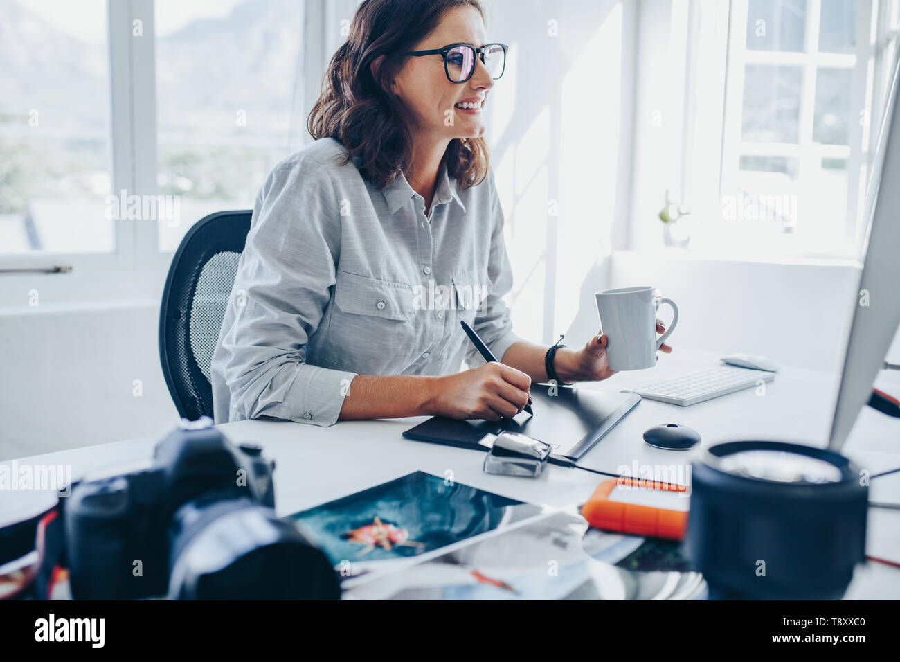Young woman sitting at desk editing images on computer. Female photographer retouching photos in office using graphic tablet and digital pen. Stock Photo