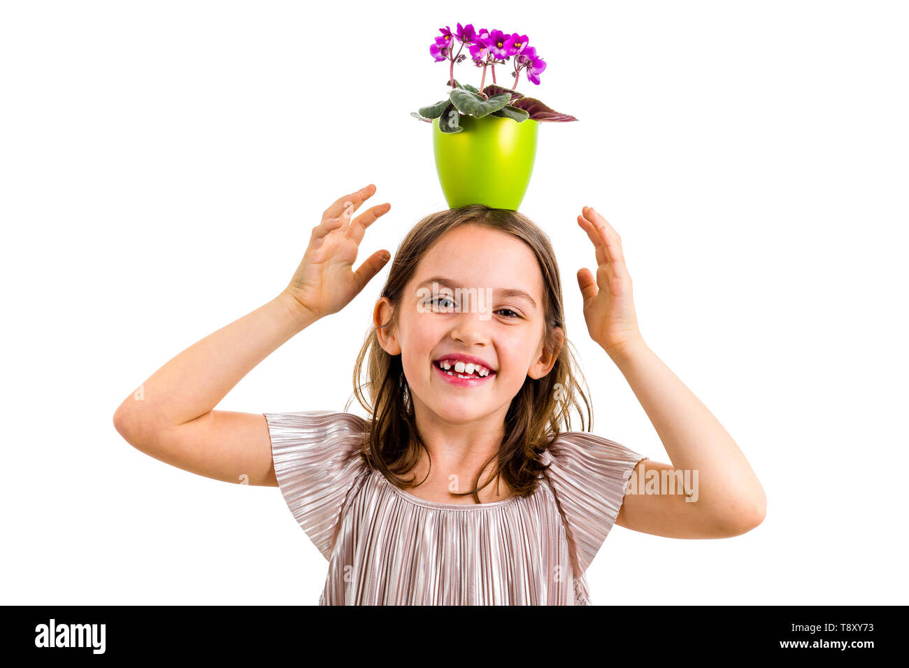 Girl carrying flower pot on head, having fun smiling. Little girl in dress holding green flower pot with viola flowers on her head having fun, goofing Stock Photo