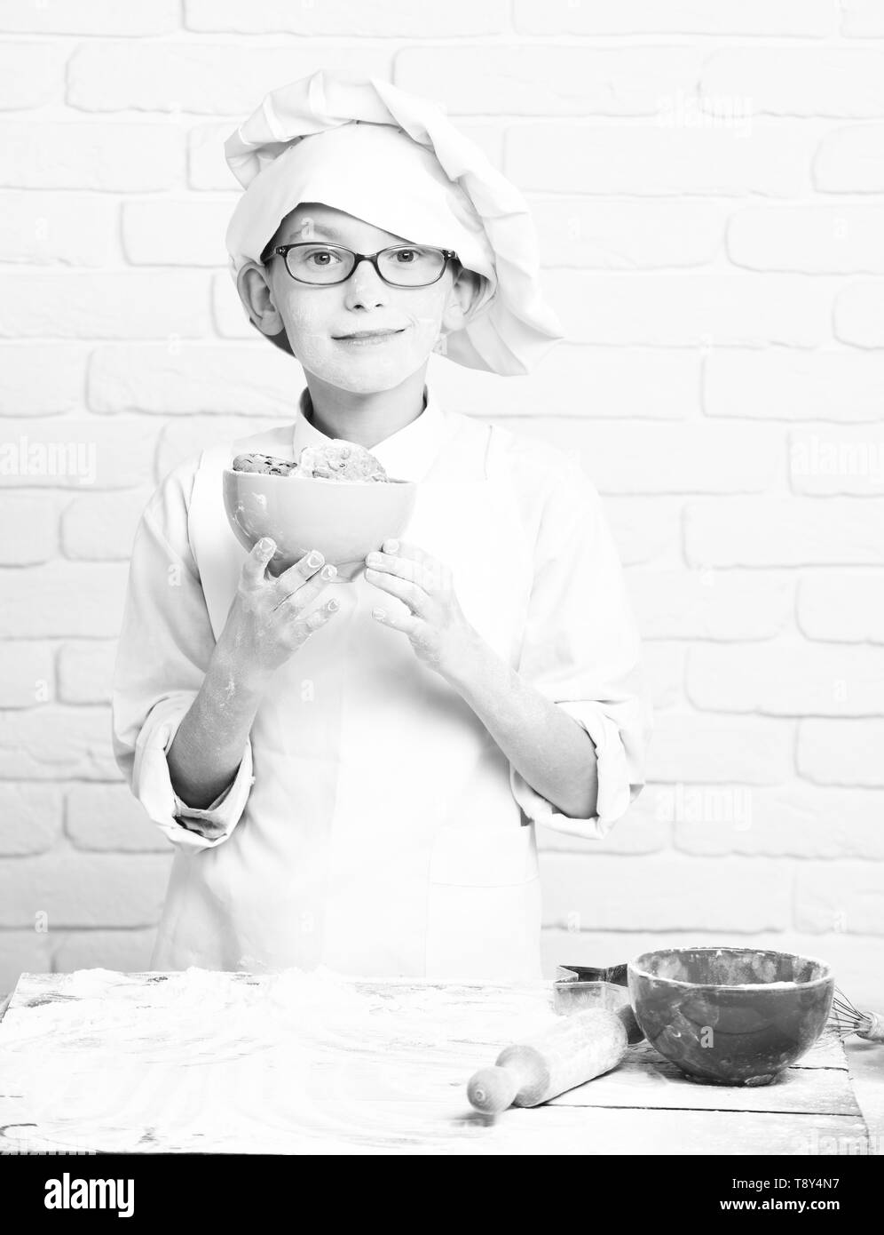 young boy cute cook chef in white uniform and hat on stained face flour with glasses standing near table with rolling pin and holding turquoise bowl with chocolate cookies on brick wall background. Stock Photo