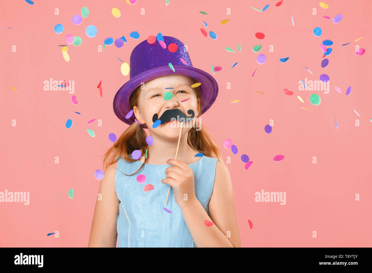 Happy father's day. Funny little girl with fake mustache and wearing a hat under falling confetti on a pink background. Family concept Stock Photo