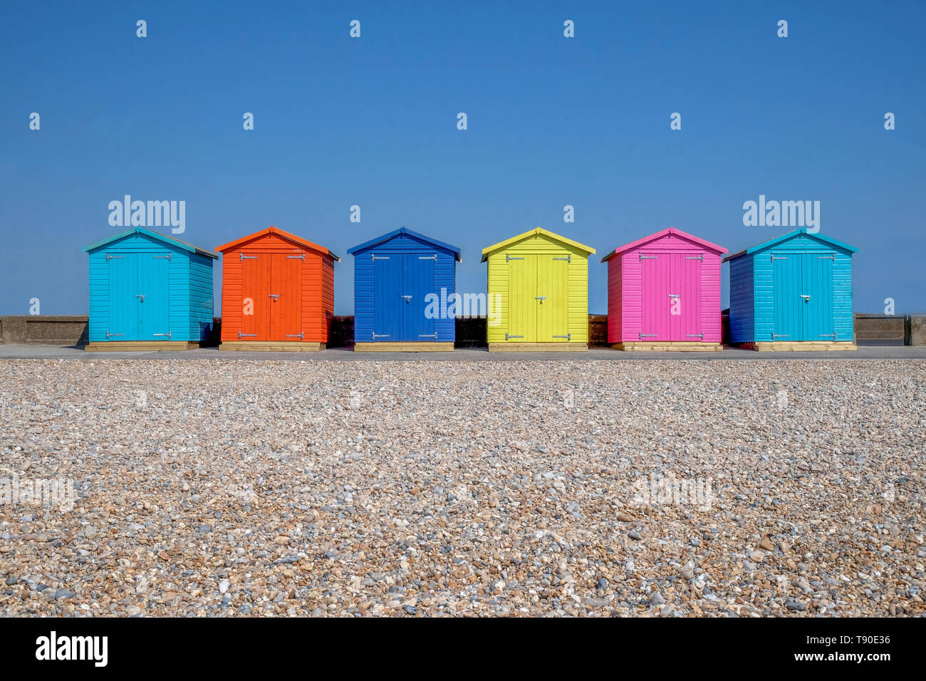 A line of six brightly coloured Beach huts, each one is a different colour in the foreground is a pebble beach and behind is a bright blue sky, Seafor Stock Photo