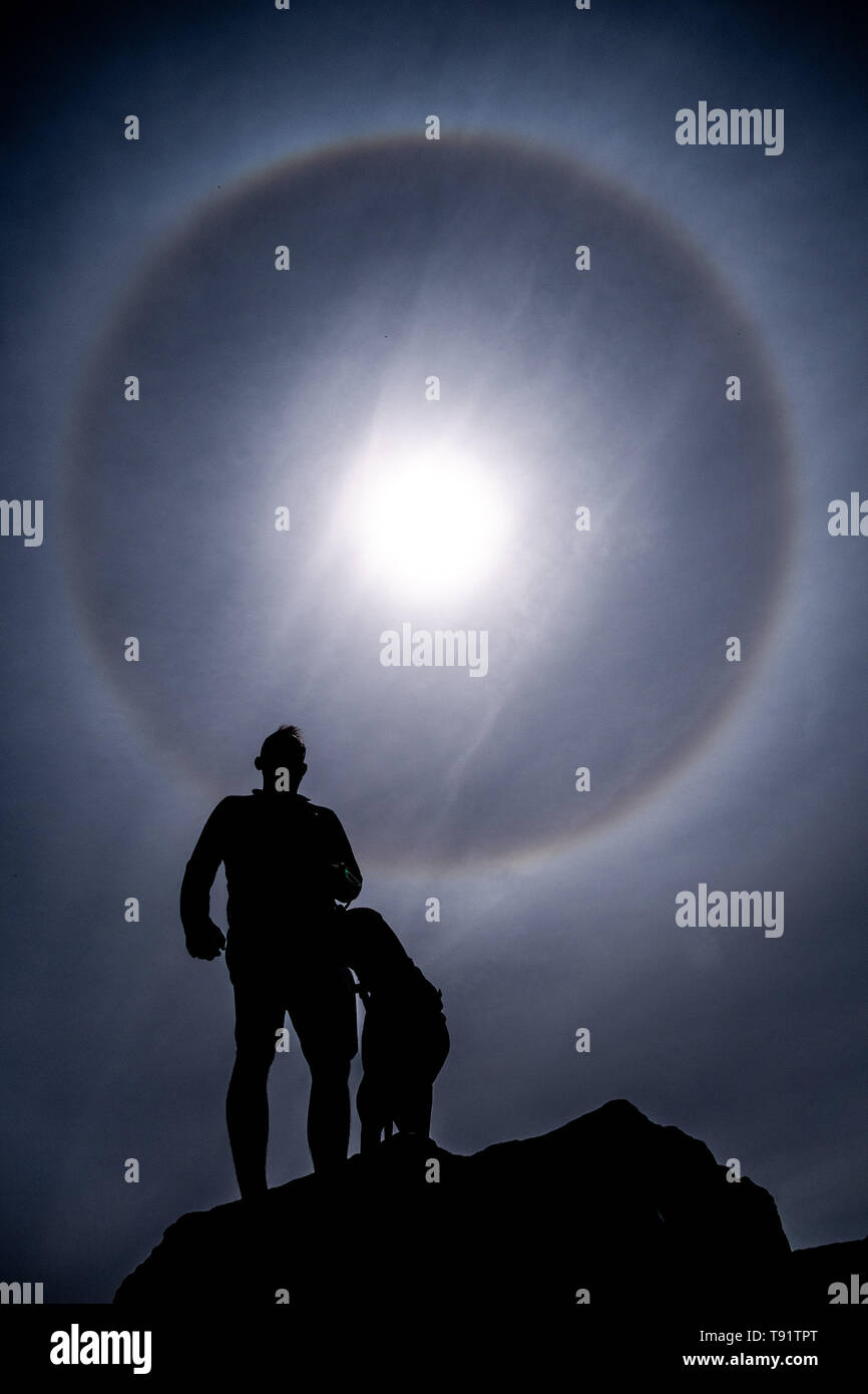 Aberystwyth Wales UK, Thursday 16 May 2019  UK Weather: A man and his dog arr silhouetted as perfect circular sun halo , formed as the rays of the sun shine through ice crystals in the upper atmosphere,  fills the sky above Aberystwyth. The halos, always at 22º from the sun,  often  indicate that rain will fall within the next 24 hours, since the cirrostratus clouds that cause them can signify an approaching frontal system. The  weather is set to change overnight from the fine warm conditions to cooler and wetter days, more typical of mid May.  photo credit Keith Morris / Alamy Live News Stock Photo