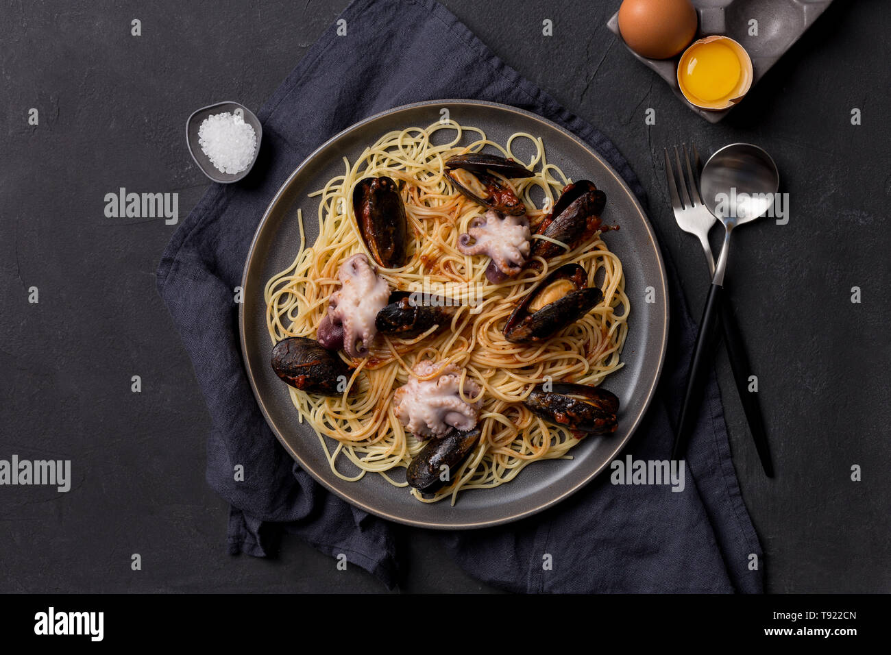 Tasty pasta with octopus and mussels on gray plate with tableware, napkin, salt and chicken egg on gray background Stock Photo