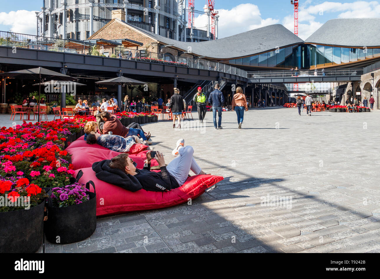 People lounging on red cushions by a bed of bright red flowers in the recently opened public space at Coal Drops Yard, King's Cross, London, UK, 2019 Stock Photo