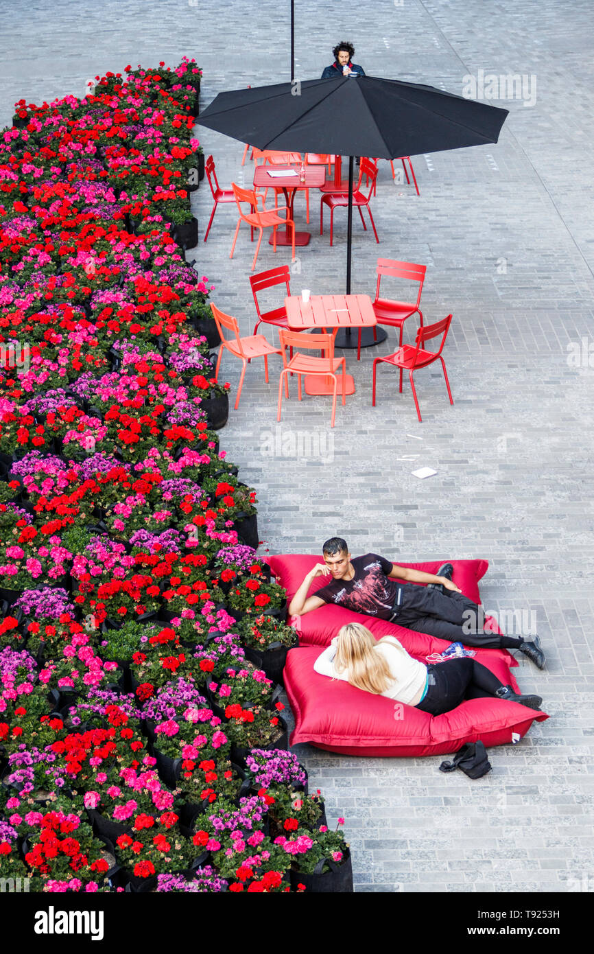 A couple lounging on red cushions by a swathe of bright red geraniums in the recently opened public space at Coal Drops Yard, King's Cross, London, UK Stock Photo
