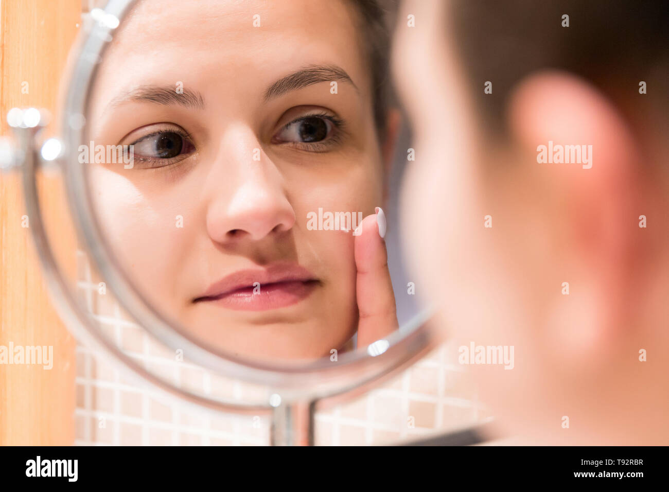 Young girl in front of a bathroom mirror putting cream on a red pimple