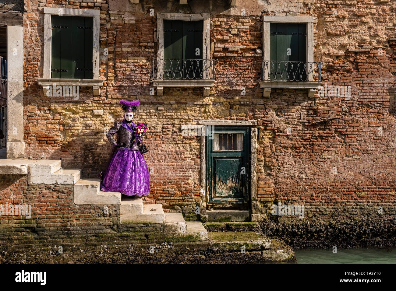A feminin masked person in a beautiful creative costume, posing in front of an ailing house facade across a small water canal, celebrating the Venetia Stock Photo