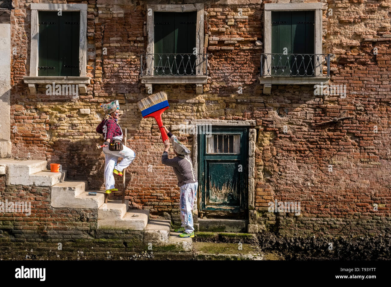 Two as painters and craftsmen masked persons in beautiful creative costumes, posing in front of an ailing house facade across a small water canal, cel Stock Photo