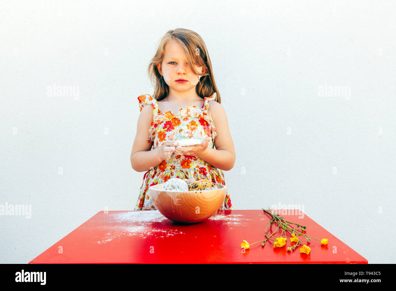 portrait of a little girl with a soiled face flour, isolated on white background, wearing a flower theme dress. A bowl with cookies and yellow flowers Stock Photo