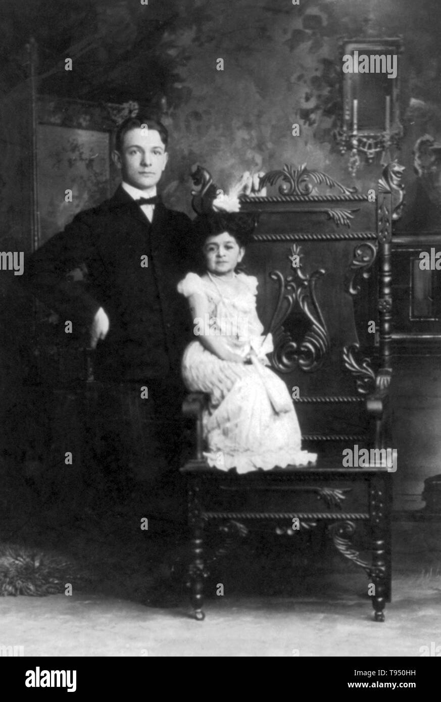 Entitled: 'Mrs. Anthony C. Woeckener of Erie, Pennsylvania, better known as Chiquita, The Doll Queen' seated on arm of chair, with a normal size young man standing alongside. Chiquita, The Doll Queen was born in either Cuba or Mexico. She was an entertainer with the traveling Bostock-Ferari Carnival. Some suspect her 1901 marriage at age 31 to 17 year old Woeckener was done for publicity. Chiquita was a hit at the Pan-American Exposition in 1901. Stock Photo