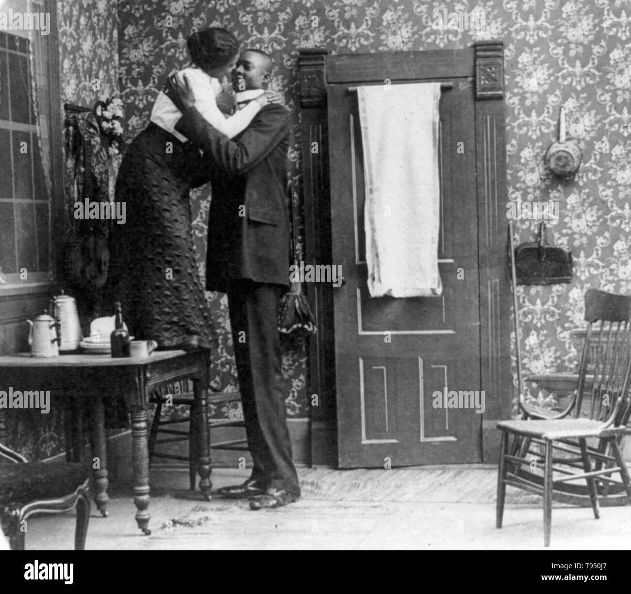 Unidentified African-American woman standing on table and embracing a tall African-American man. Gigantism, also known as giantism, is a condition characterized by excessive growth and height significantly above average. In humans, this condition is caused by over-production of growth hormone in childhood resulting in people between 7 feet and 9 feet in height. Stock Photo