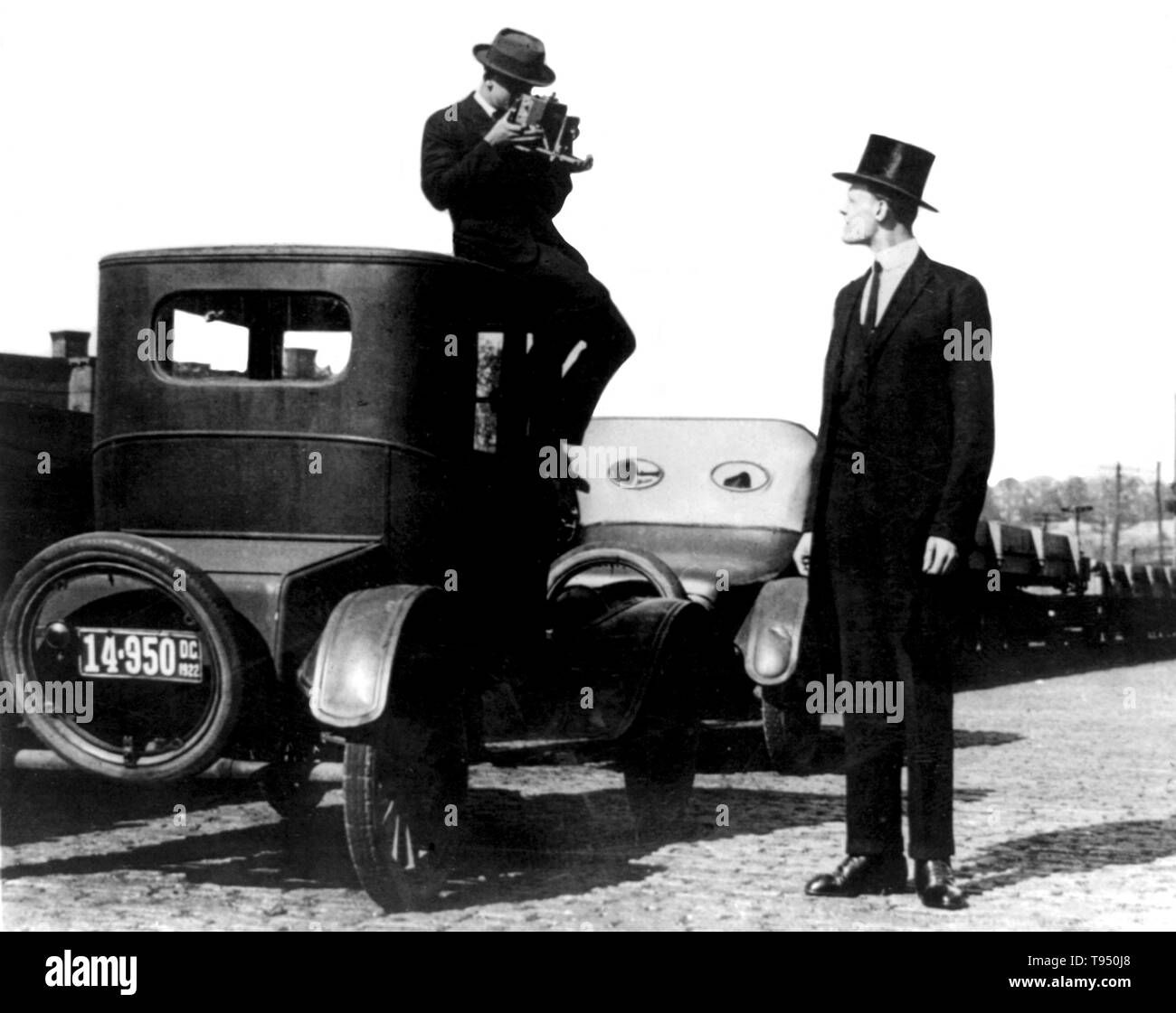 Entitled: 'Photographer seated on top of car photographing tall man in top hat.' Gigantism, also known as giantism, is a condition characterized by excessive growth and height significantly above average. In humans, this condition is caused by over-production of growth hormone in childhood resulting in people between 7 feet and 9 feet in height. National Photo Company, 1922. Stock Photo