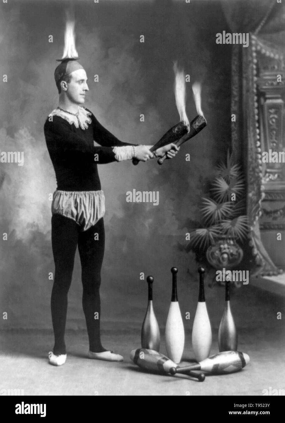 Entitled: 'J.T. Doyle holding 2 burning juggling clubs'. Juggling is a physical skill, performed by a juggler, involving the manipulation of objects for recreation, entertainment, art or sport. The most recognizable form of juggling is toss juggling. Juggling can be the manipulation of one object or many objects at the same time, using one or many hands. Jugglers often refer to the objects they juggle as props. The most common props are balls, clubs, or rings. Some jugglers use more dramatic objects such as knives, fire torches or chainsaws. Photographed by J.E. Pasonault, 1902. Stock Photo