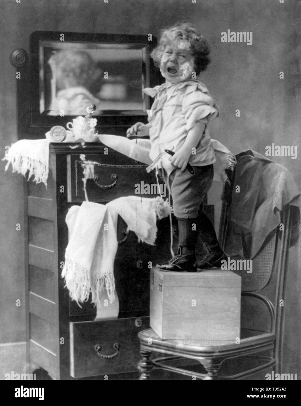 Entitled: 'Crying child with bleeding hand standing on box on chair in front of mirror above dresser and holding razor with soap on face'. Photographed by E. Donald Roberts, 1898. Stock Photo