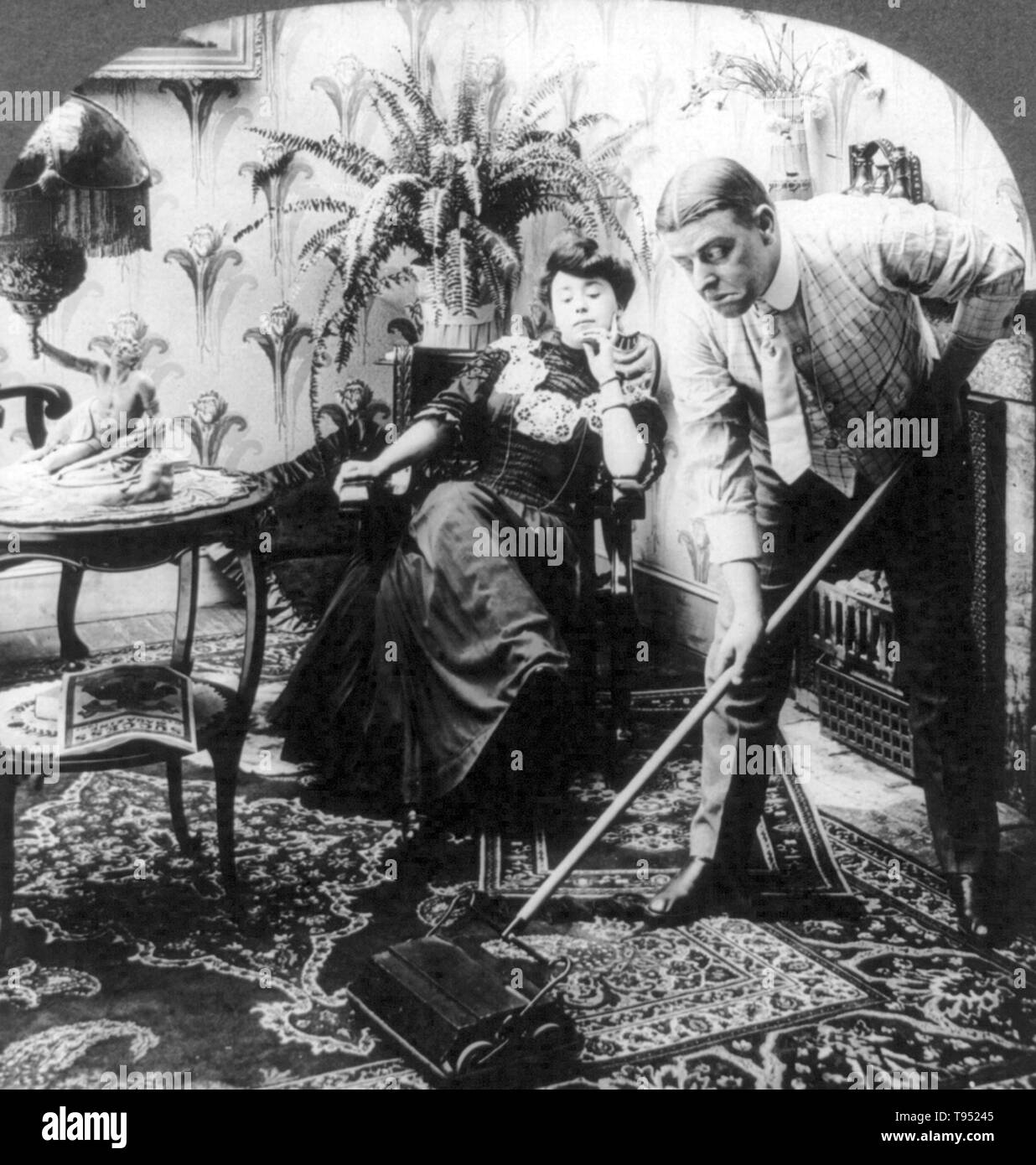 Entitled: 'Take your time Mr. Peck - haste makes waste' shows a man using carpet sweeper as woman sits and watches. Cropped stereograph photographed by E.W. Kelley October 14, 1907. Stock Photo