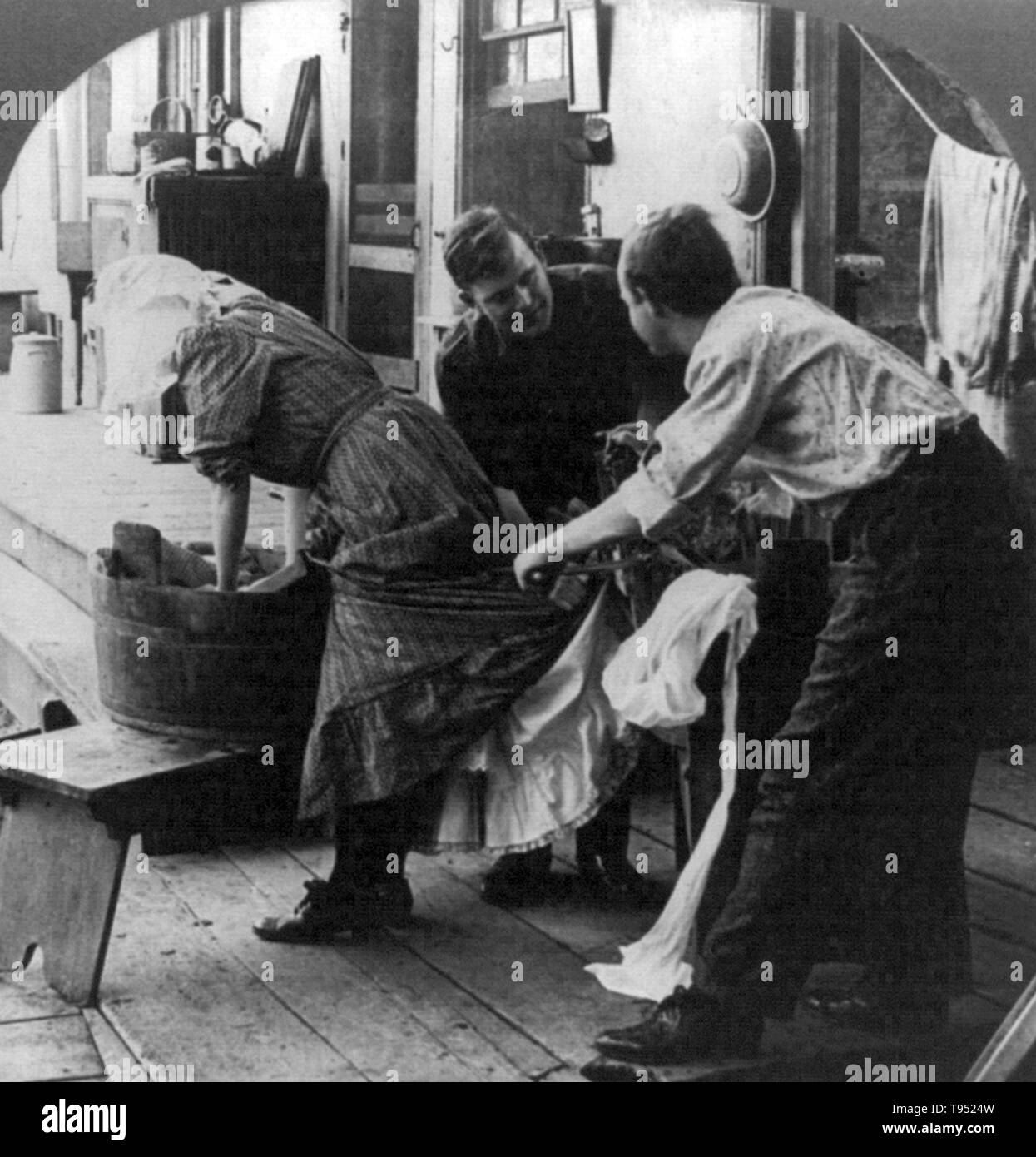 Entitled: 'Wringer up George, quick!' show a woman doing laundry in tub, as two men put bottom of her dress and skirt through wringer. Cropped stereograph photographed by E.W. Kelley, 1906. Stock Photo