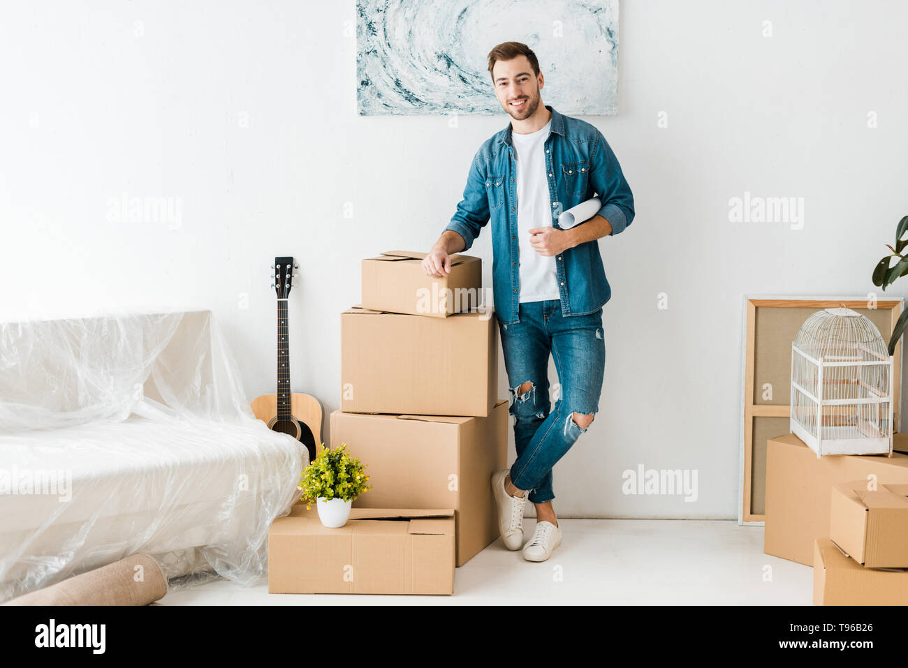 full length view of smiling man in jeans standing near cardboard boxes at new home Stock Photo