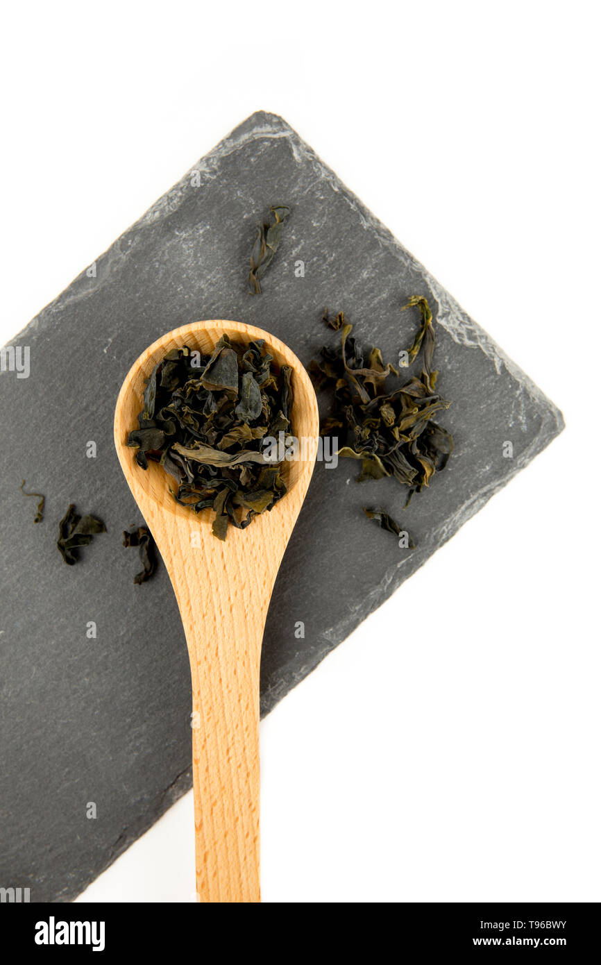 Small pieces of dried Wakame (Undaria pinnatifida), edible seaweed, ingredient to different foods in wooden spoon on black stone cutting board isolate Stock Photo