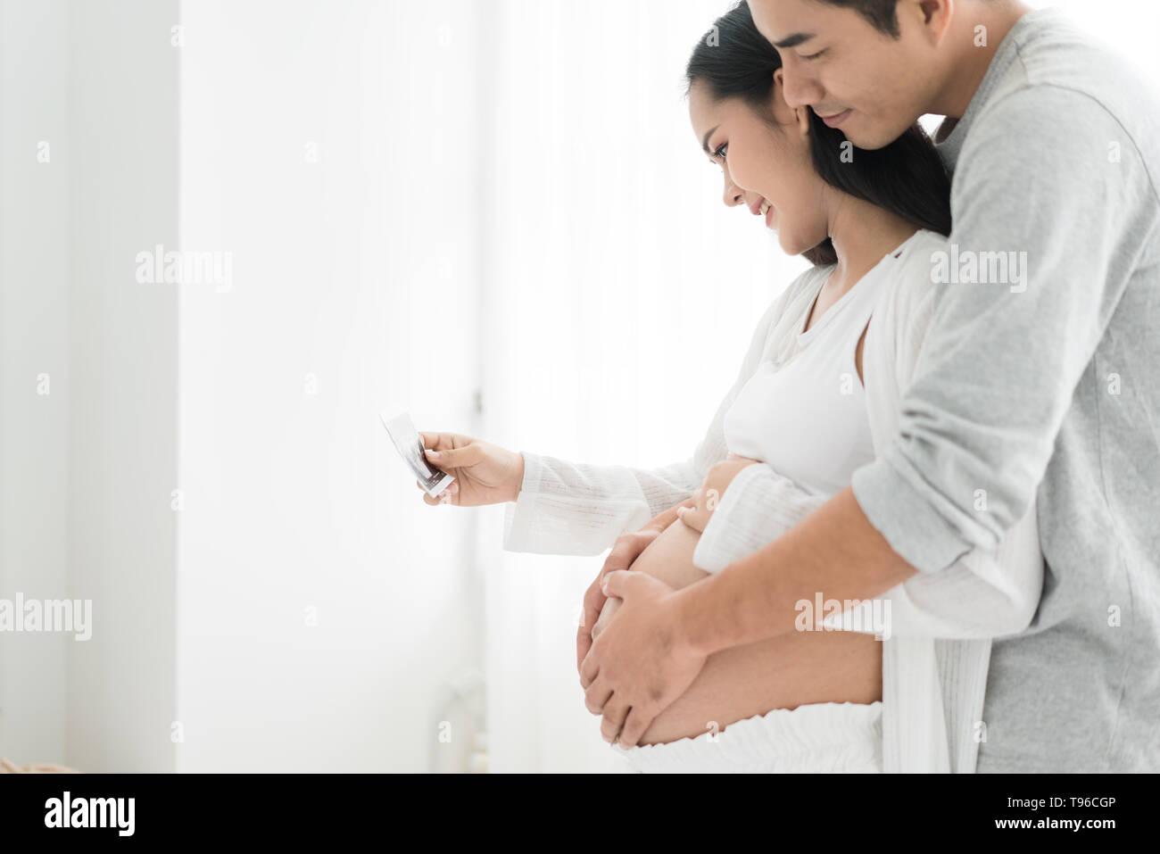 Beautiful Asian pregnant woman and Asian handsome husband holding and looking a sonogram in house. Concept of pregnancy, health care, gynecology, medi Stock Photo