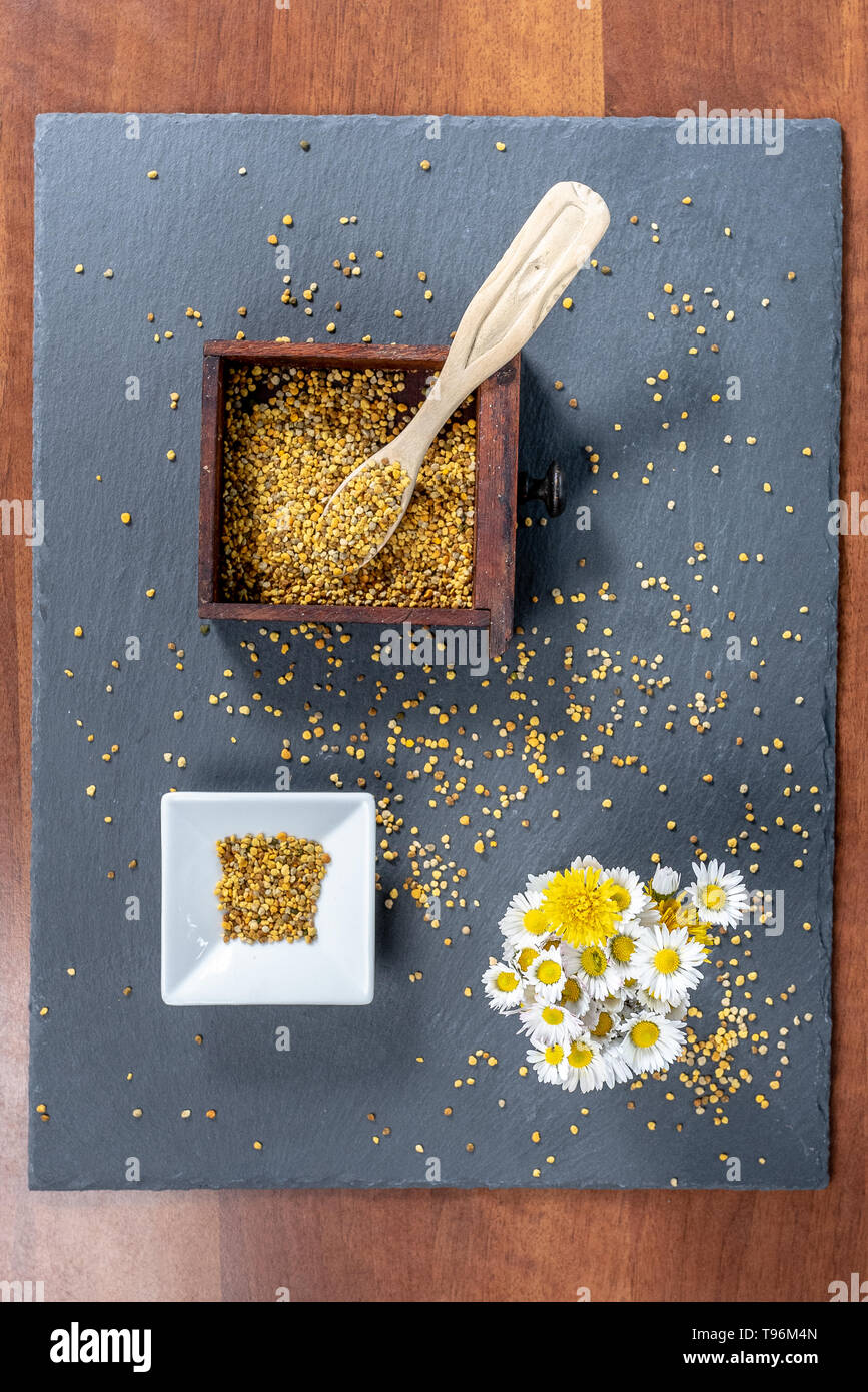 photo composed of a square bowl of white ceramic, a wooden box full of pollen grains of bees and a bunch of daisies, on a slate board, top view Stock Photo