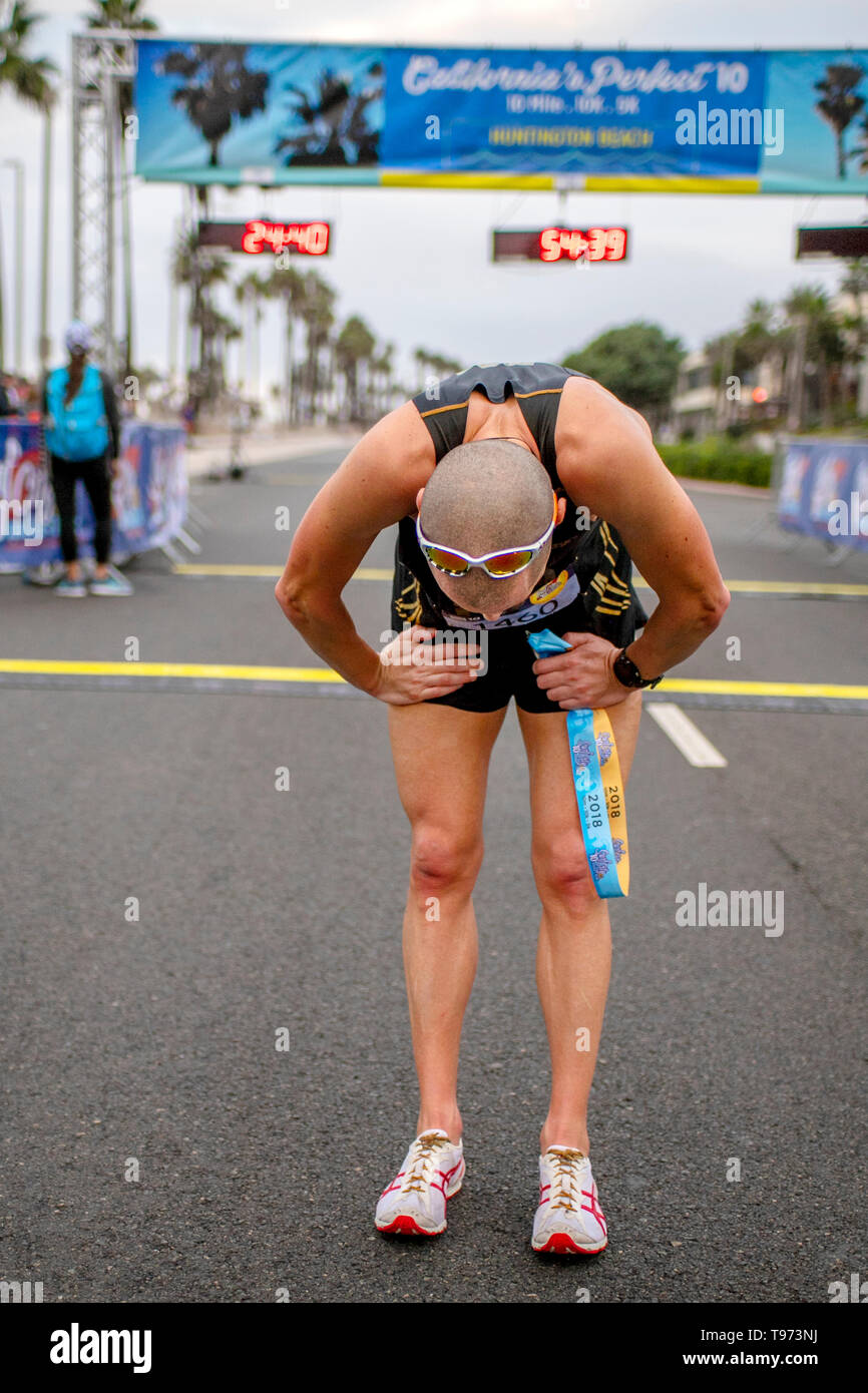 Exhausted after crossing the finish line, a 10-mile competitive runner bends over to catch his breath as he holds a ribbon signifying he finish the race in Huntington Beach, CA. Stock Photo