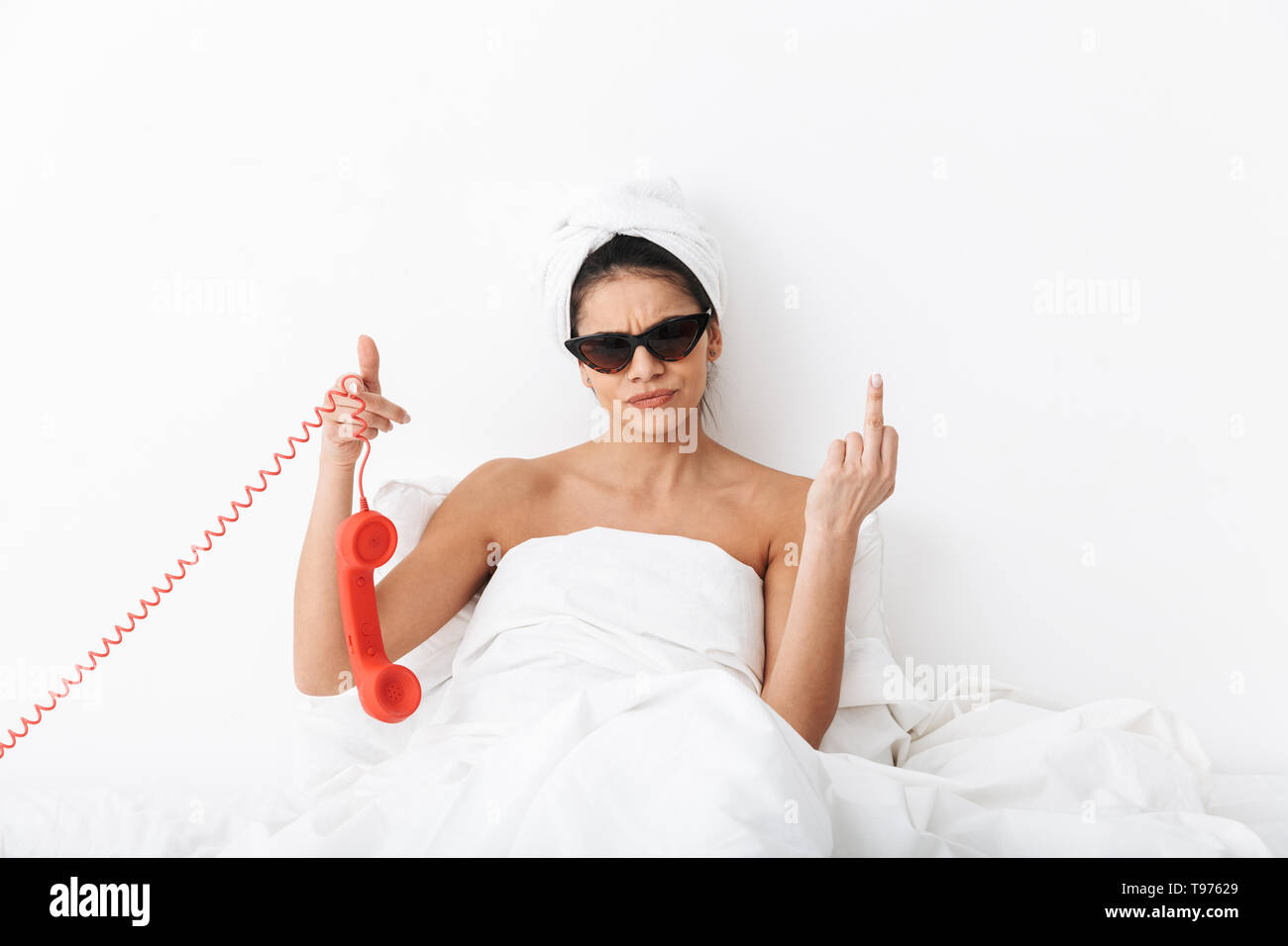 Upset young woman sitting in bed after shower wrapped in blanket, wearing sunglasses, talking on a landline phone Stock Photo