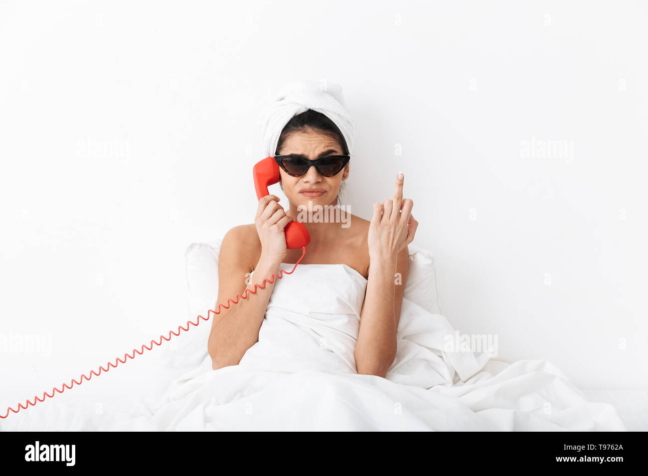 Upset young woman sitting in bed after shower wrapped in blanket, wearing sunglasses, talking on a landline phone Stock Photo