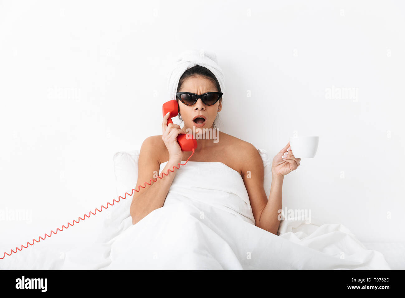 Upset young woman sitting in bed after shower wrapped in blanket, wearing sunglasses, talking on a landline phone, holding a cup Stock Photo
