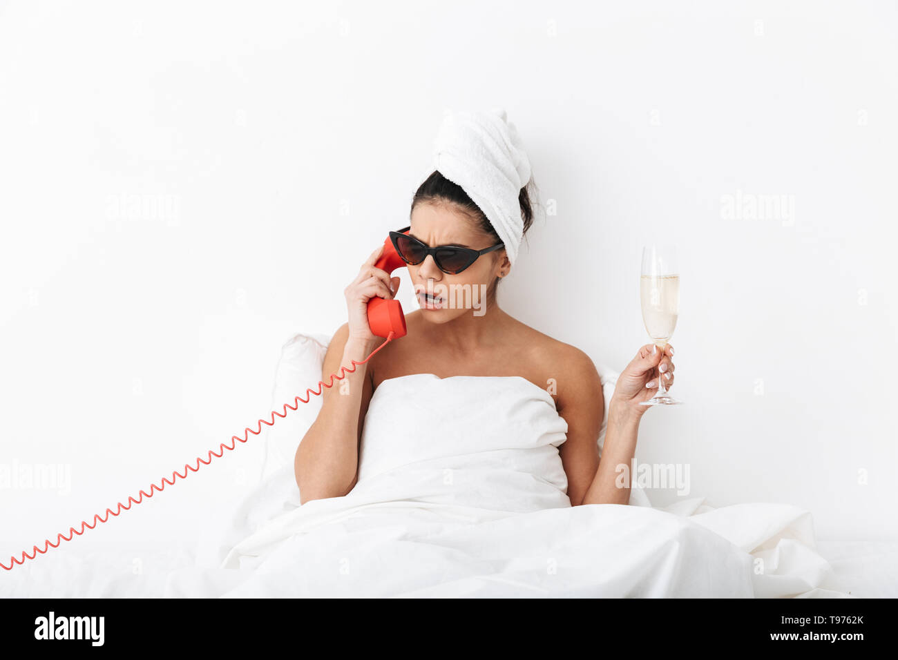 Upset young woman sitting in bed after shower wrapped in blanket, wearing sunglasses, talking on a landline phone, holding glass of champagne Stock Photo
