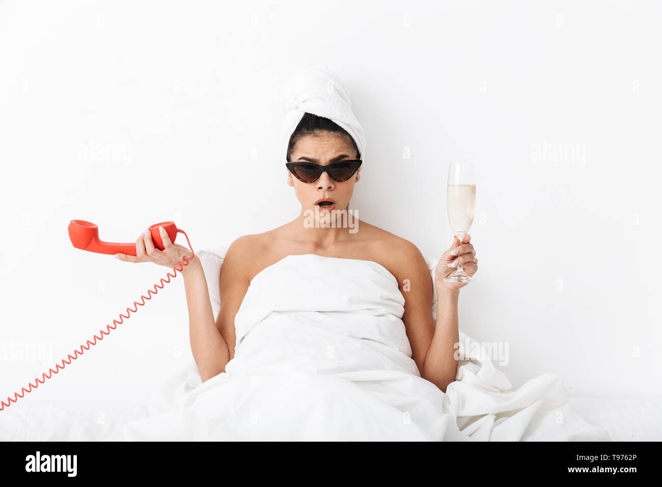 Cheerful young woman sitting in bed after shower wrapped in blanket, wearing sunglasses, talking on a landline phone, holding glass of champagne Stock Photo