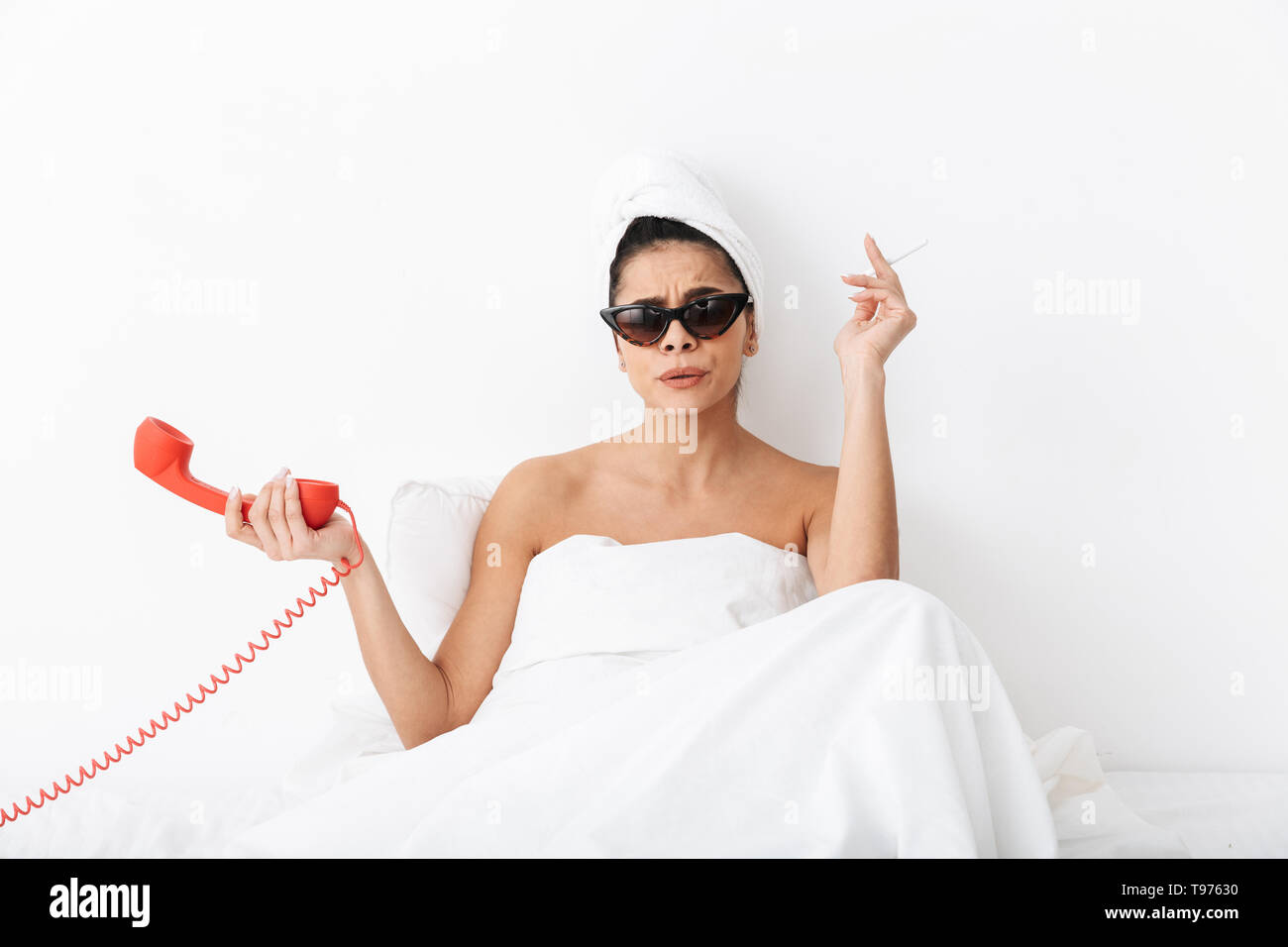 Cheerful young woman sitting in bed after shower wrapped in blanket, wearing sunglasses, smoking, talking on a landline phone Stock Photo