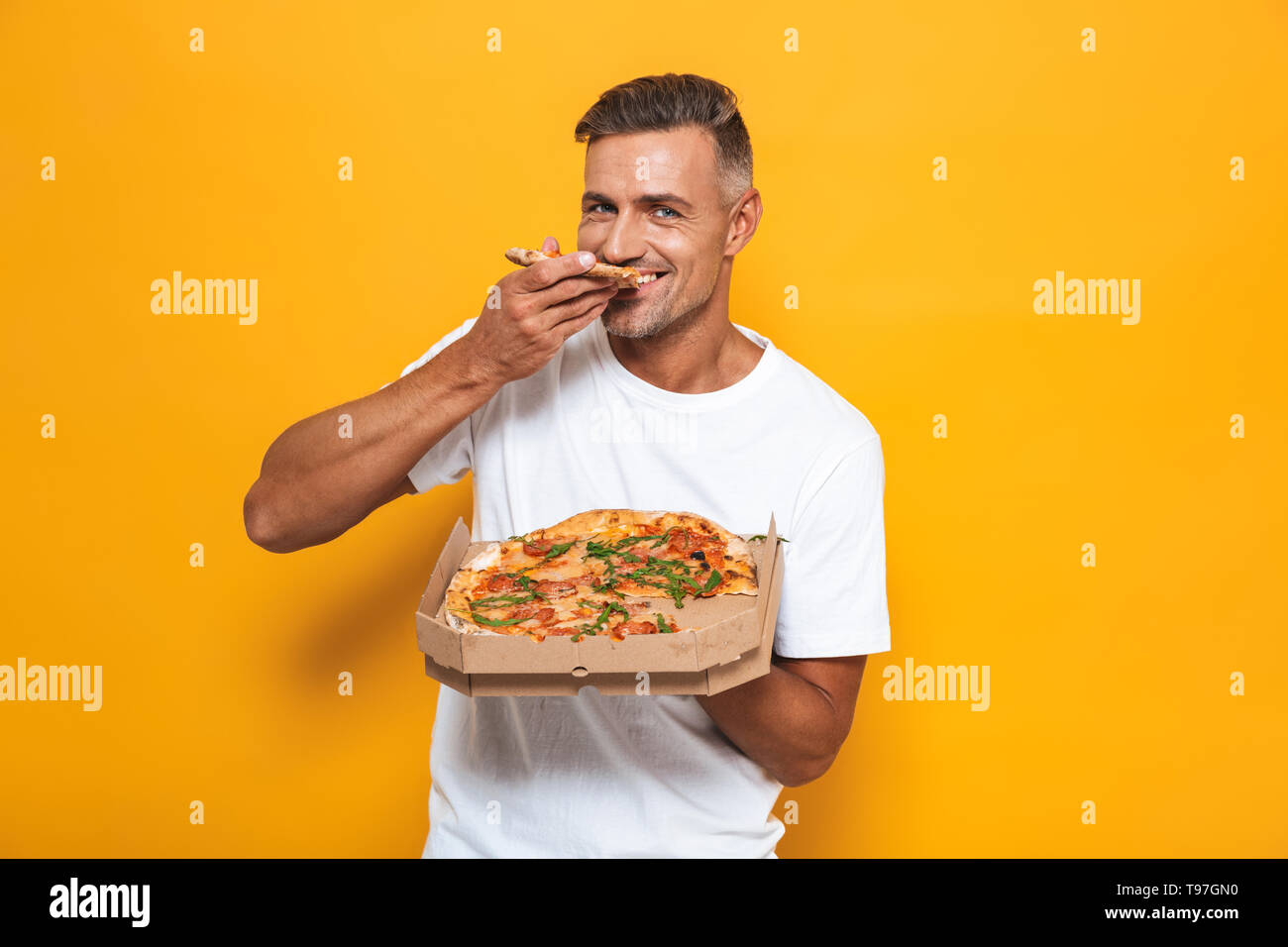 Image of joyful man 30s in white t-shirt holding and eating pizza while sta...