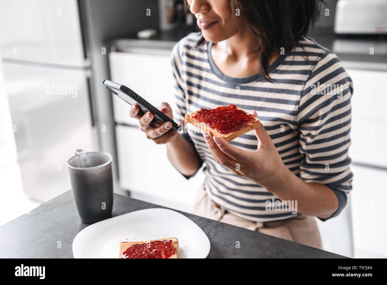 Cropped image of a young african woman having breakfast while sitting at the kitchen, using mobile phone Stock Photo