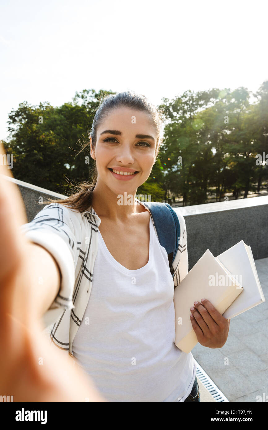 Cheerful young student girl carrying backpack outdoors, holding books