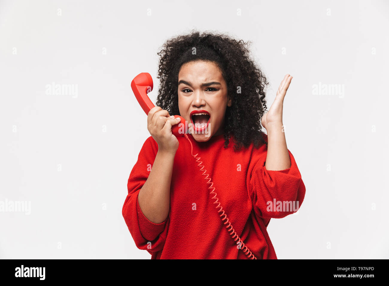 Portrait of an angry african woman standing isolated over white background, talking on a landline phone Stock Photo