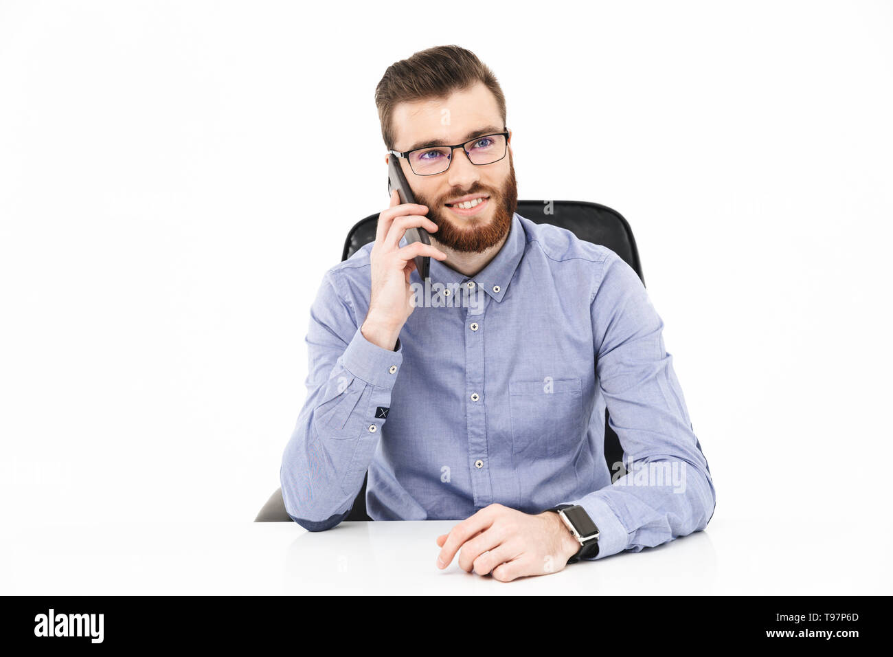 Smiling bearded elegant man in eyeglasses talking by smartphone and looking away while sitting by the table over grey background Stock Photo